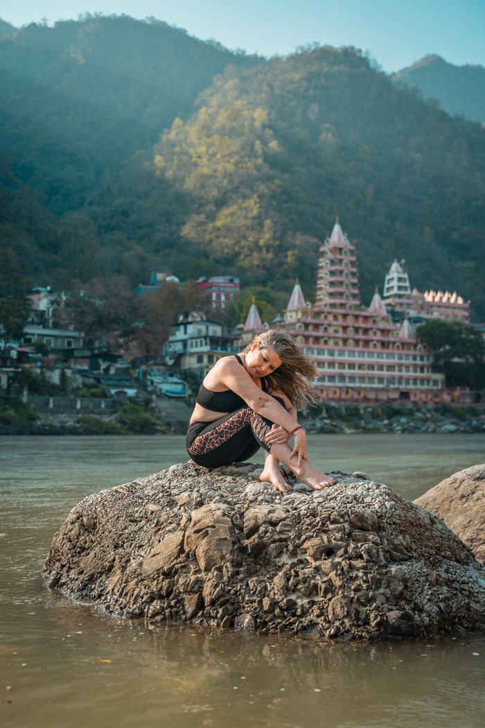 A woman meditates on a rock by the river with a scenic backdrop of mountains and temples.