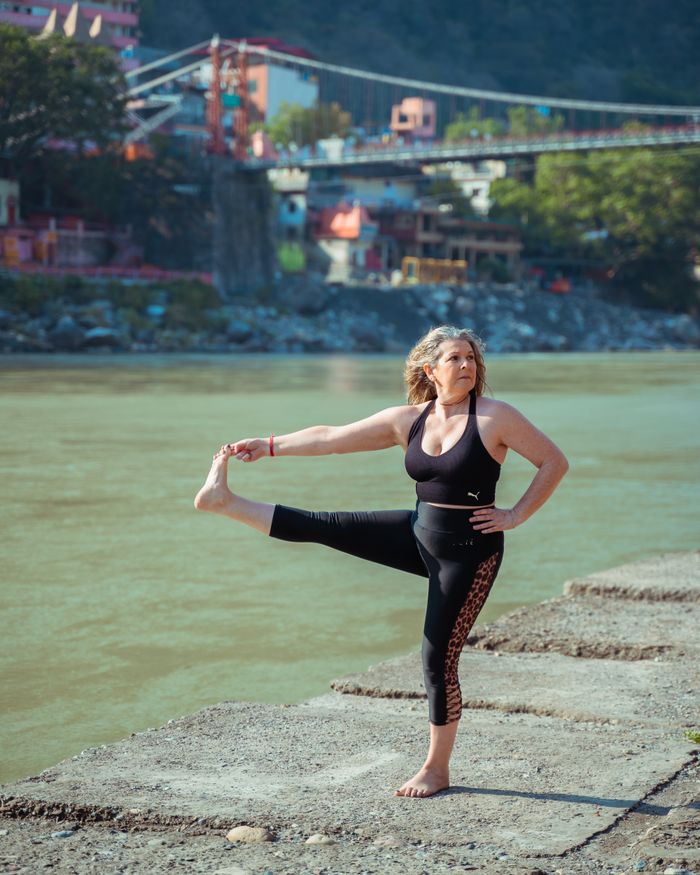 Woman practicing yoga pose near river with scenic background, embodying fitness and wellness.