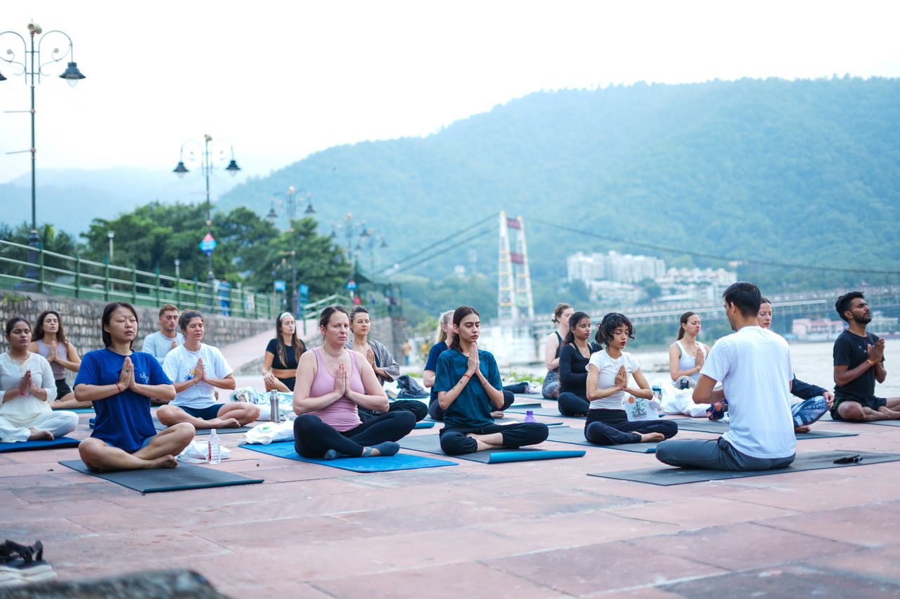 about-02 A diverse group practicing yoga by the river in Rishikesh, India, surrounded by scenic mountains.